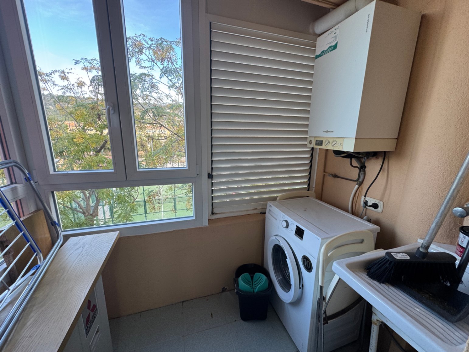 Zona de lavadero funcional con lavadora, caldera de gas y fregadero. El espacio cuenta con amplios ventanales que aportan luz natural y vistas al entorno arbolado.
