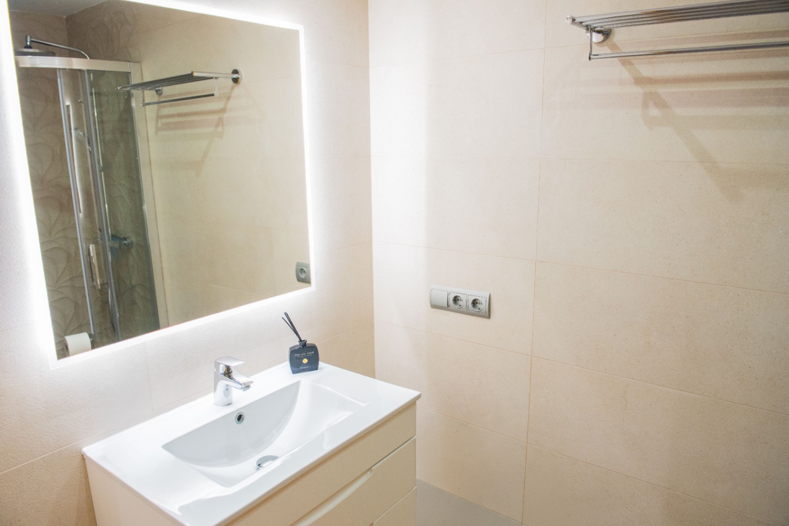 Baño moderno con lavabo blanco integrado, espejo con iluminación LED perimetral y paredes revestidas en gres cerámico de tono crema.
