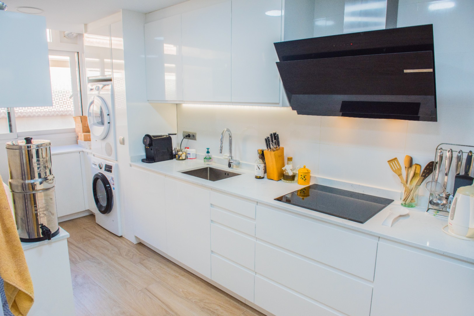 Cocina blanca minimalista con armarios de alto brillo, placa de inducción, campana negra y zona de lavandería integrada con electrodomésticos en columna.
