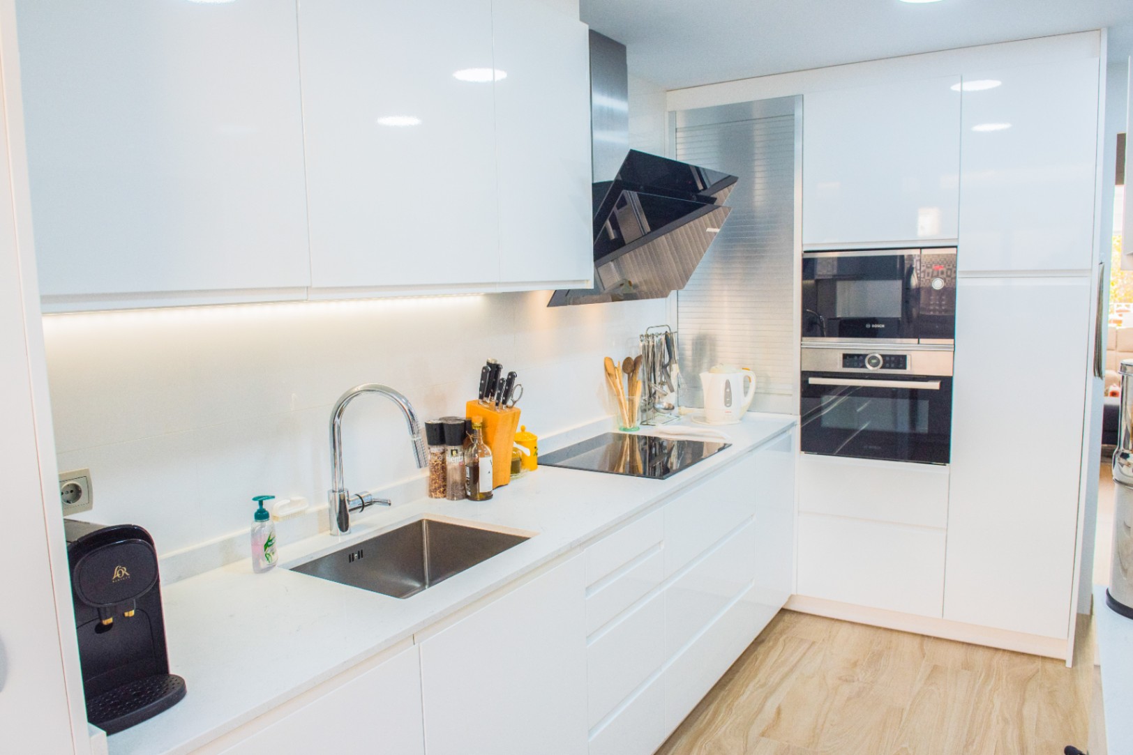 Cocina moderna con armarios blancos de alto brillo, electrodomésticos Bosch integrados y encimera de cuarzo blanco con fregadero de acero inoxidable bajo encimera.