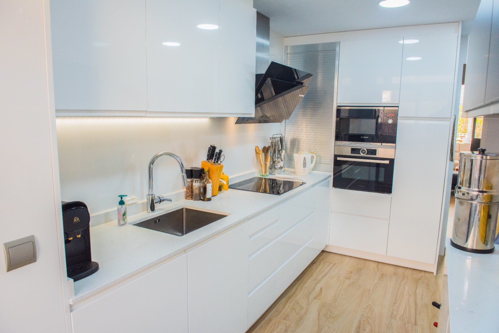 Cocina moderna con armarios blancos de alto brillo, electrodomésticos Bosch integrados y placa de inducción. Incluye encimera de cuarzo blanco y suelo de madera clara.