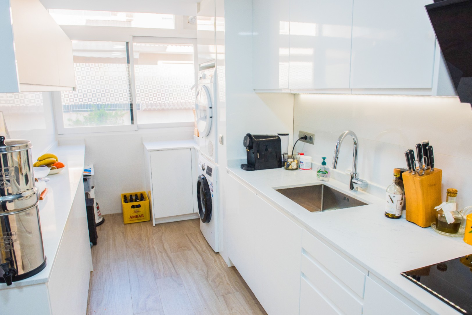 Cocina moderna de estilo galera con armarios blancos sin tiradores, zona de lavandería integrada y encimeras blancas. Incluye iluminación LED bajo los muebles y suelo de madera clara.