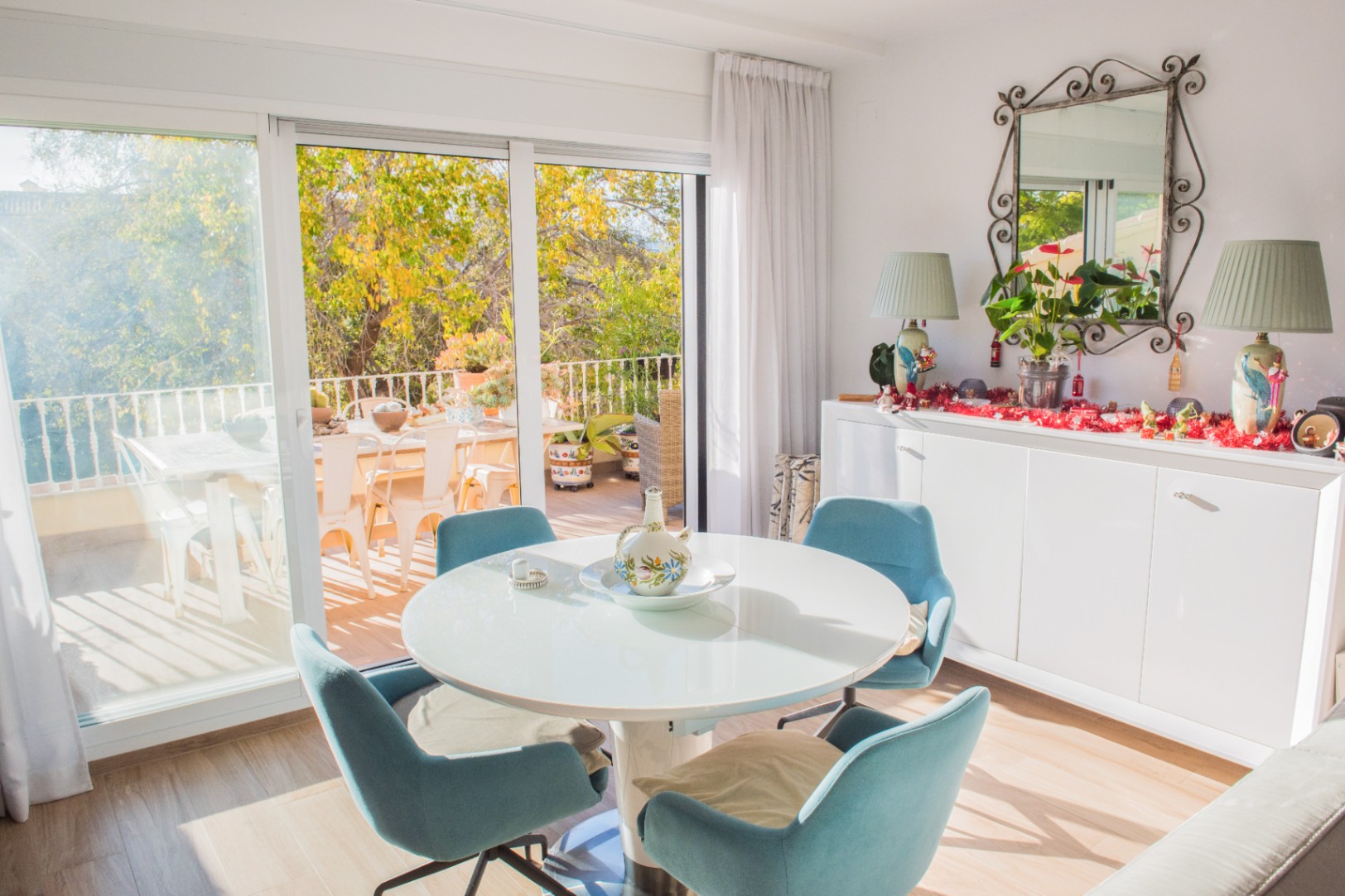 Comedor luminoso con mesa redonda blanca y sillas azules, conectado a una terraza soleada mediante grandes puertas correderas de cristal.
