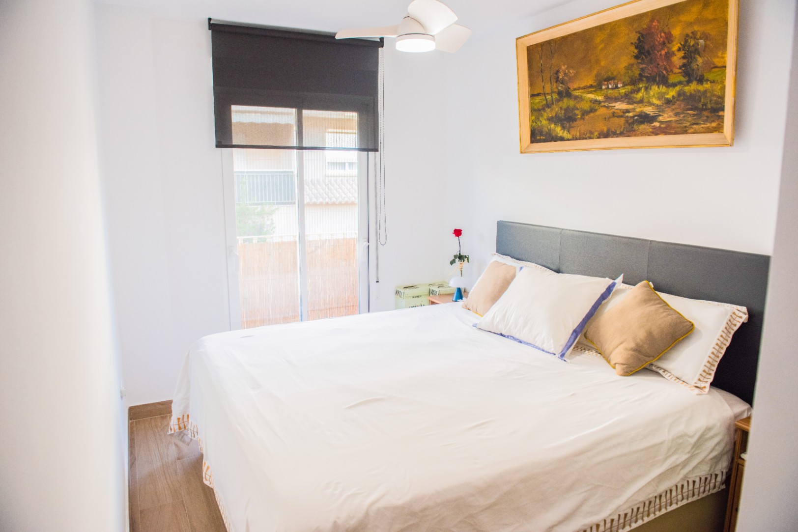 Dormitorio luminoso con cabecero gris, ventilador de techo y ventanal con estor negro. Destaca un cuadro al óleo de un paisaje sobre la cama.