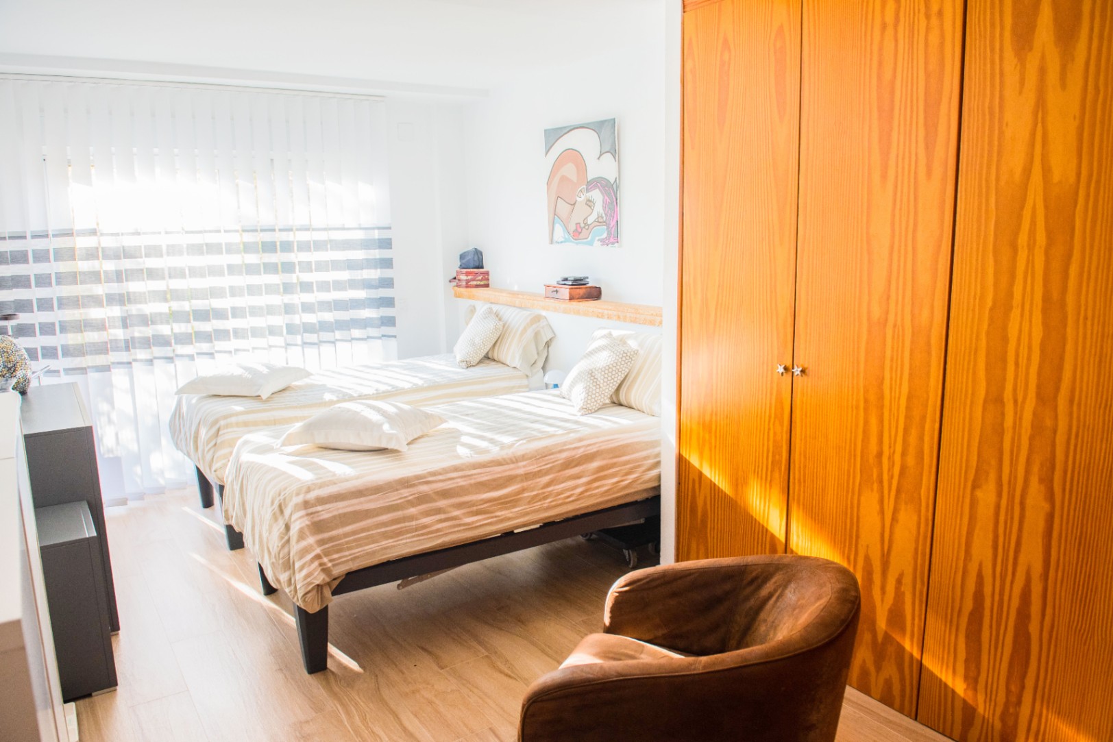 Dormitorio luminoso con dos camas individuales, armario empotrado de madera natural y un sillón de cuero sobre suelo de tarima clara.