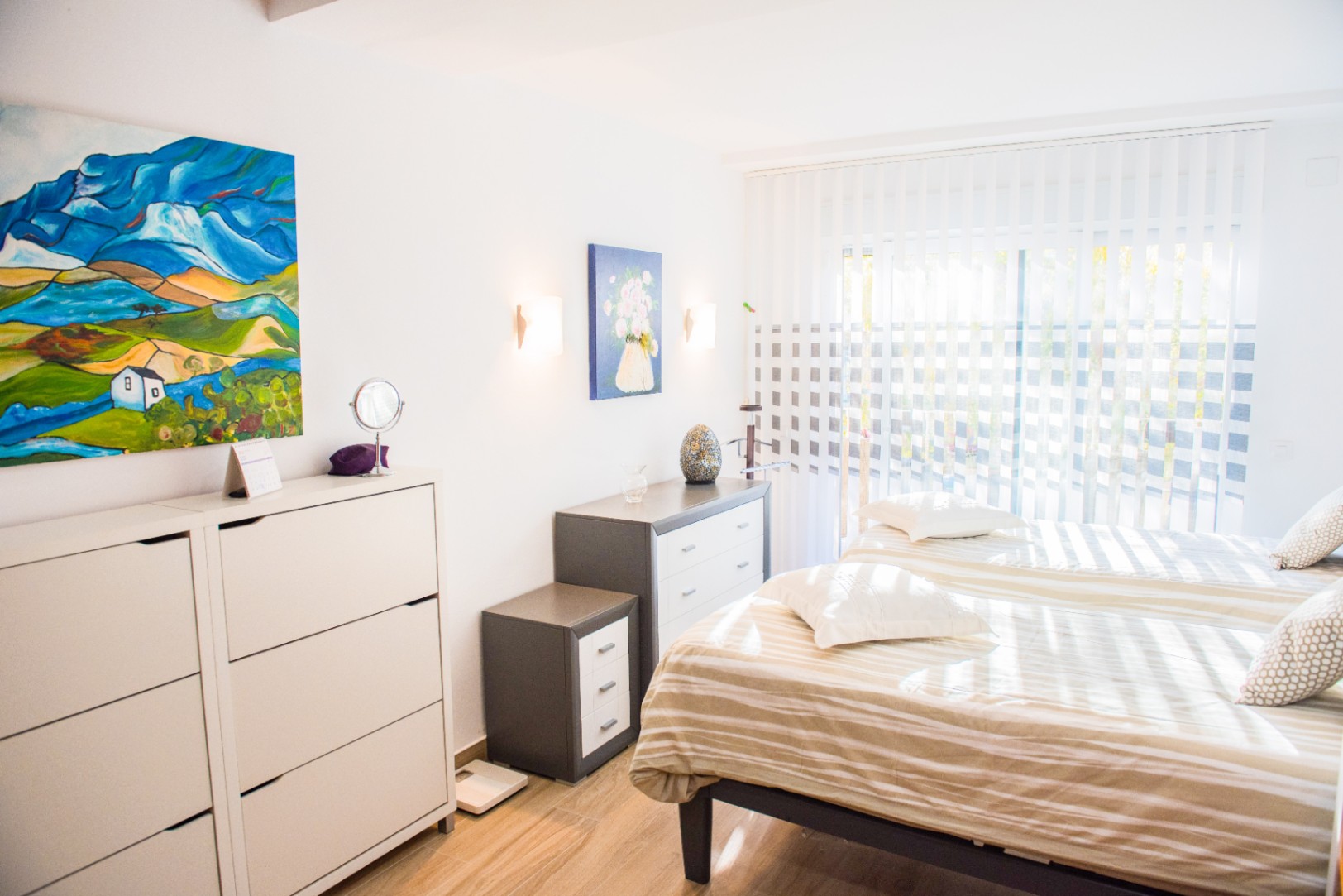 Dormitorio luminoso con dos camas individuales, cómodas de diseño minimalista y arte colorido. La luz natural entra a través de persianas verticales sobre el suelo de madera.