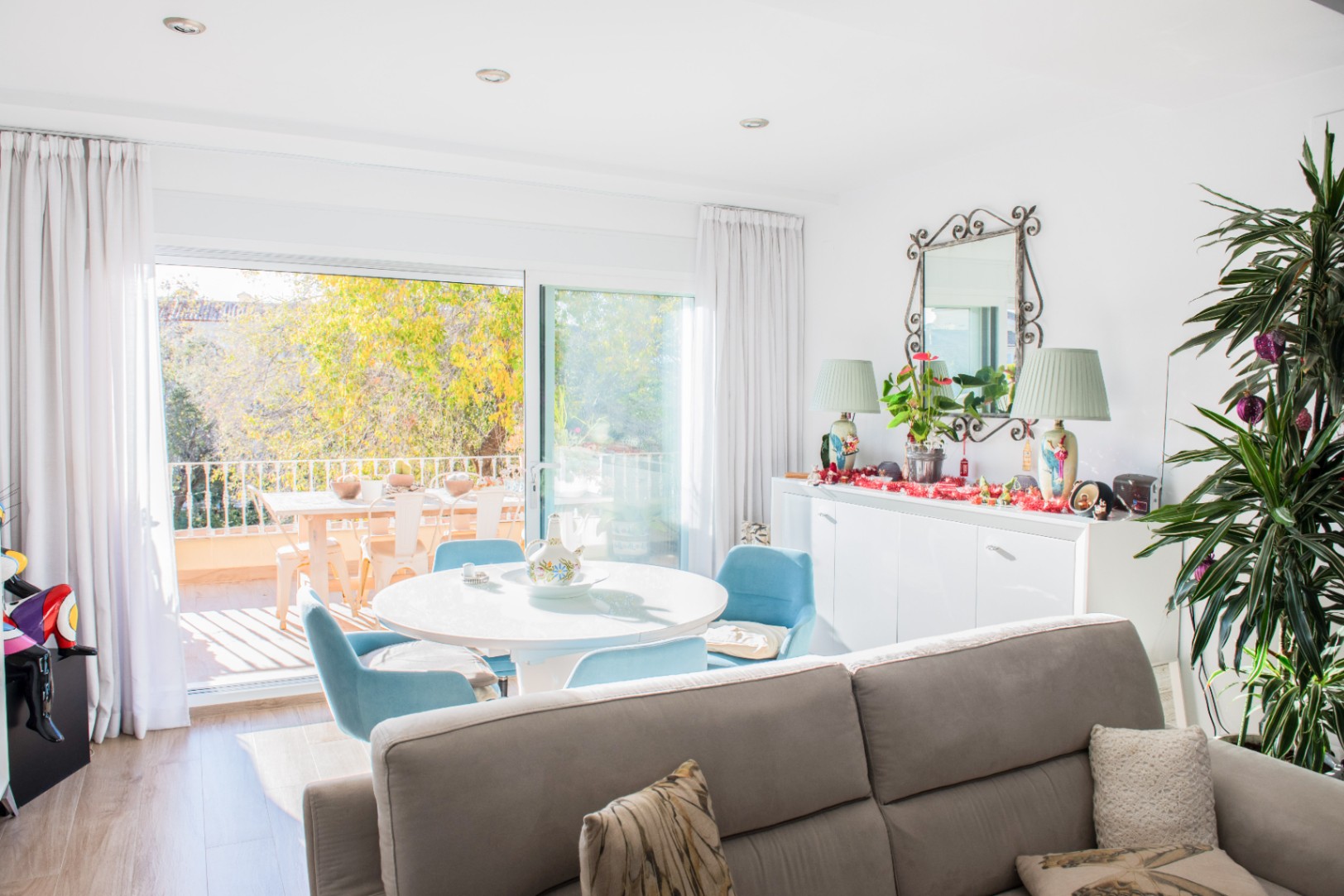 Luminosa zona de estar con mesa de comedor redonda y sillas azules, conectada a una terraza con mobiliario exterior y vistas despejadas al jardín.