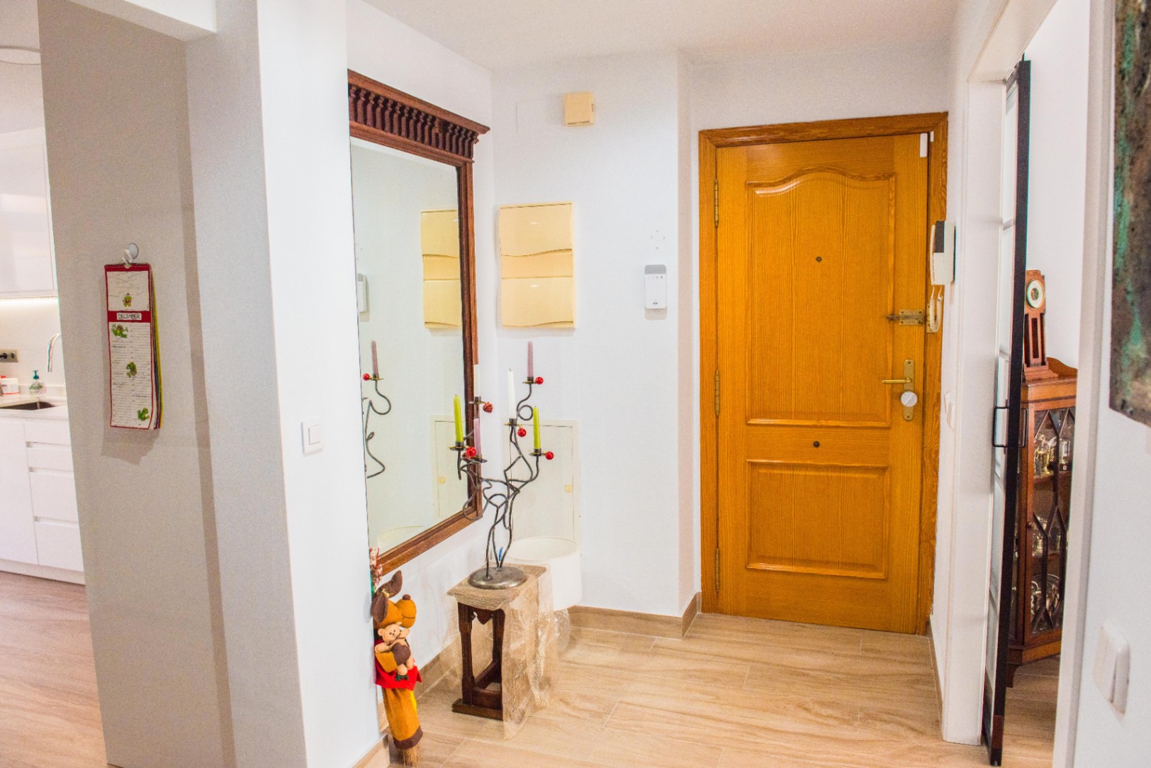 Recibidor con puerta de madera maciza, suelo de gres claro y un gran espejo con marco de madera tallada. El espacio comunica con una cocina de tonos blancos.