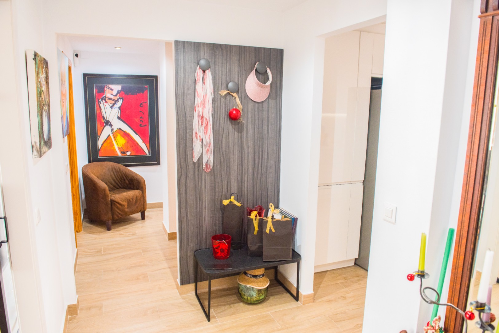 Recibidor con suelo cerámico imitación madera, panel decorativo de veta oscura con percheros y banco minimalista. Al fondo, un pasillo con arte colorido y un sillón marrón.