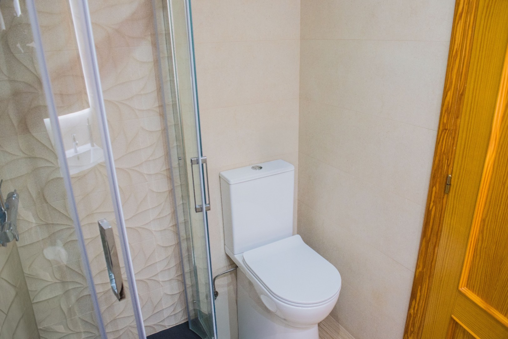 Rincón de baño con inodoro blanco y cabina de ducha de vidrio, destacando el revestimiento cerámico con relieve de hojas y grifería cromada.