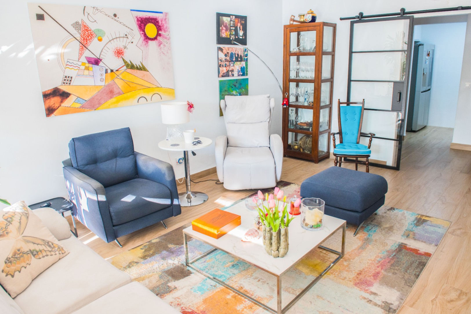 Salón con sillón azul, relax blanco y vitrina de madera. Destaca el arte abstracto, una alfombra multicolor y una puerta corredera de cristal con marco industrial negro.