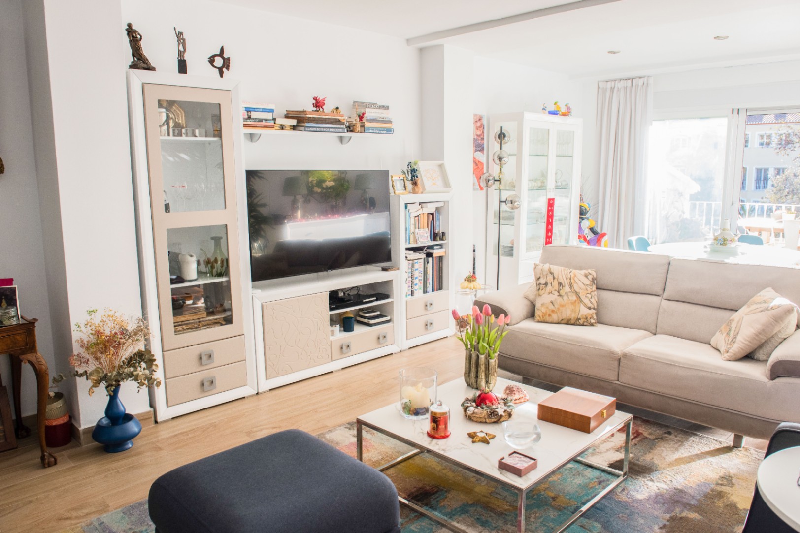 Salón luminoso con sofá de tela beige, mesa de centro de mármol y mueble modular blanco. Grandes puertas acristaladas aportan abundante luz natural al espacio.