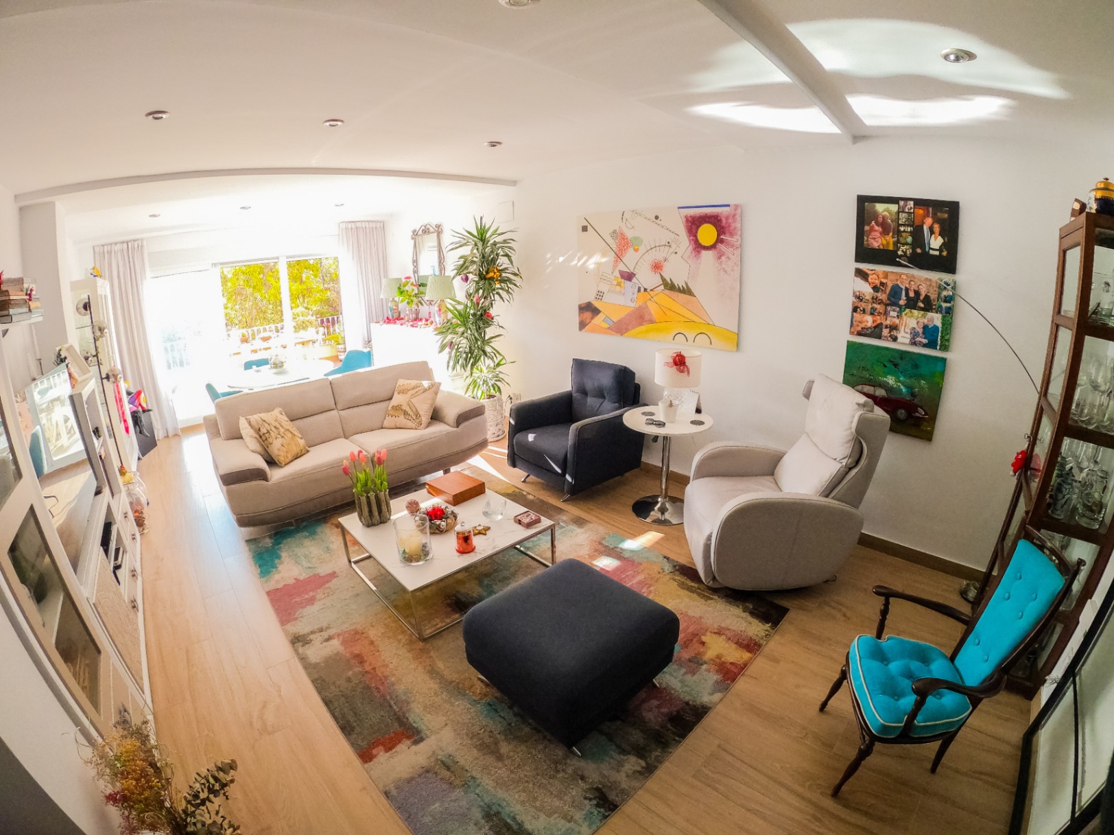 Salón luminoso con suelos de madera clara, alfombra abstracta multicolor y mobiliario variado. Grandes puertas acristaladas aportan luz natural y acceso a una terraza con vegetación.