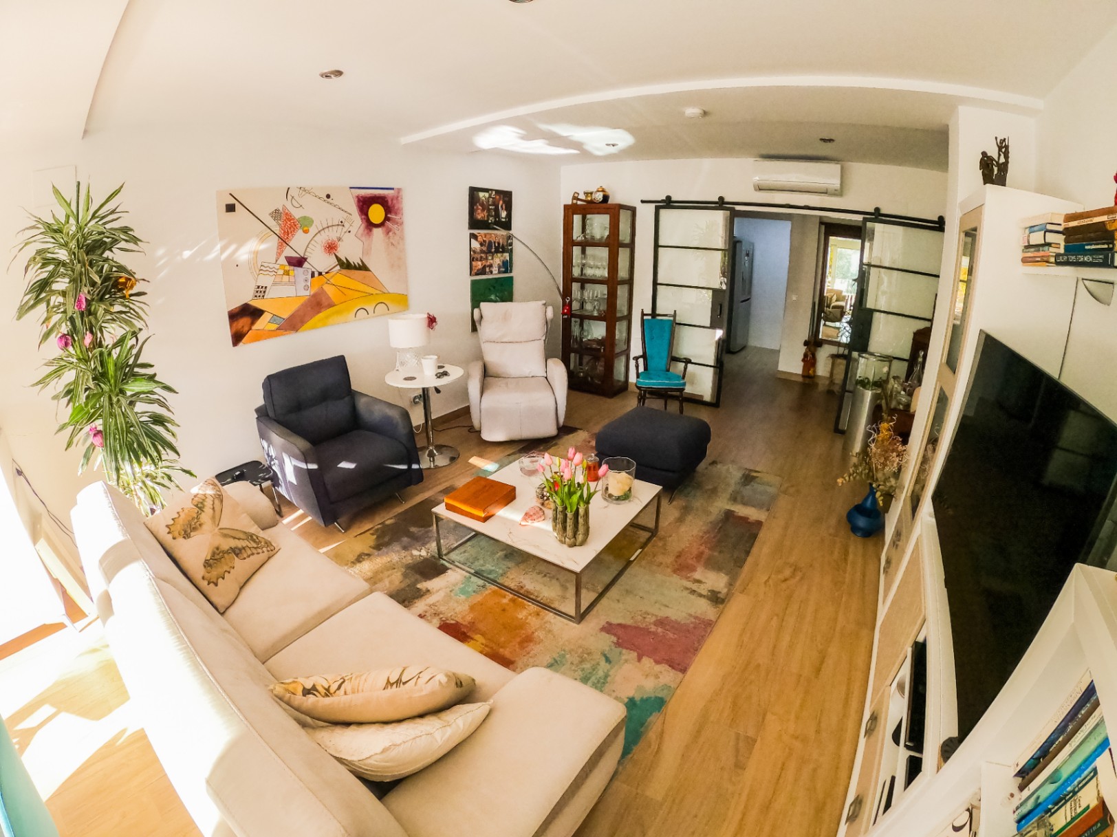 Salón moderno con sofá rinconero blanco, arte abstracto y mesa de centro de mármol sobre alfombra multicolor. Puertas correderas de estilo industrial conectan los espacios.