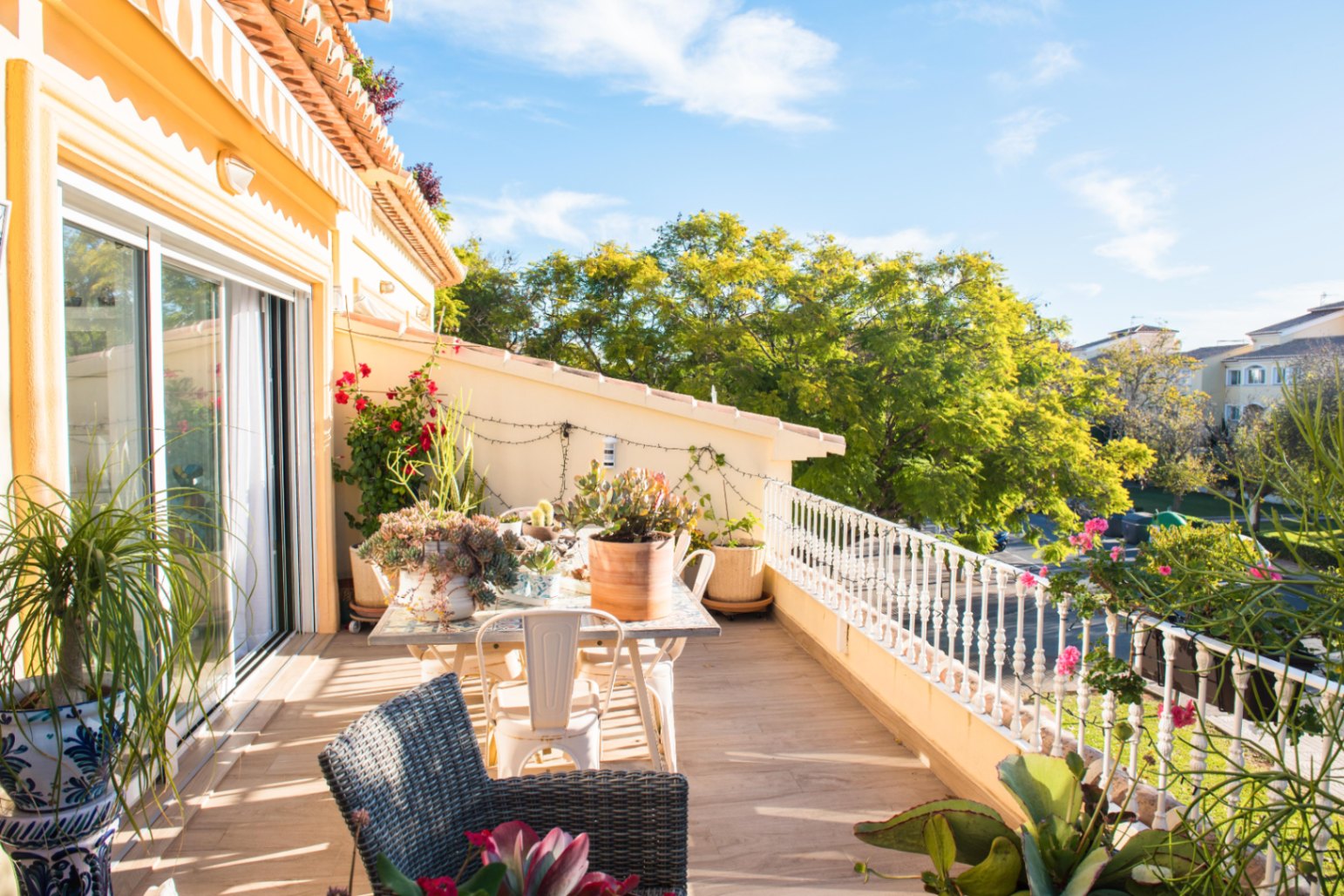 Terraza con suelo cerámico de efecto madera, zona de comedor exterior y barandilla blanca. Decorada con plantas variadas y vistas a zonas verdes arboladas.