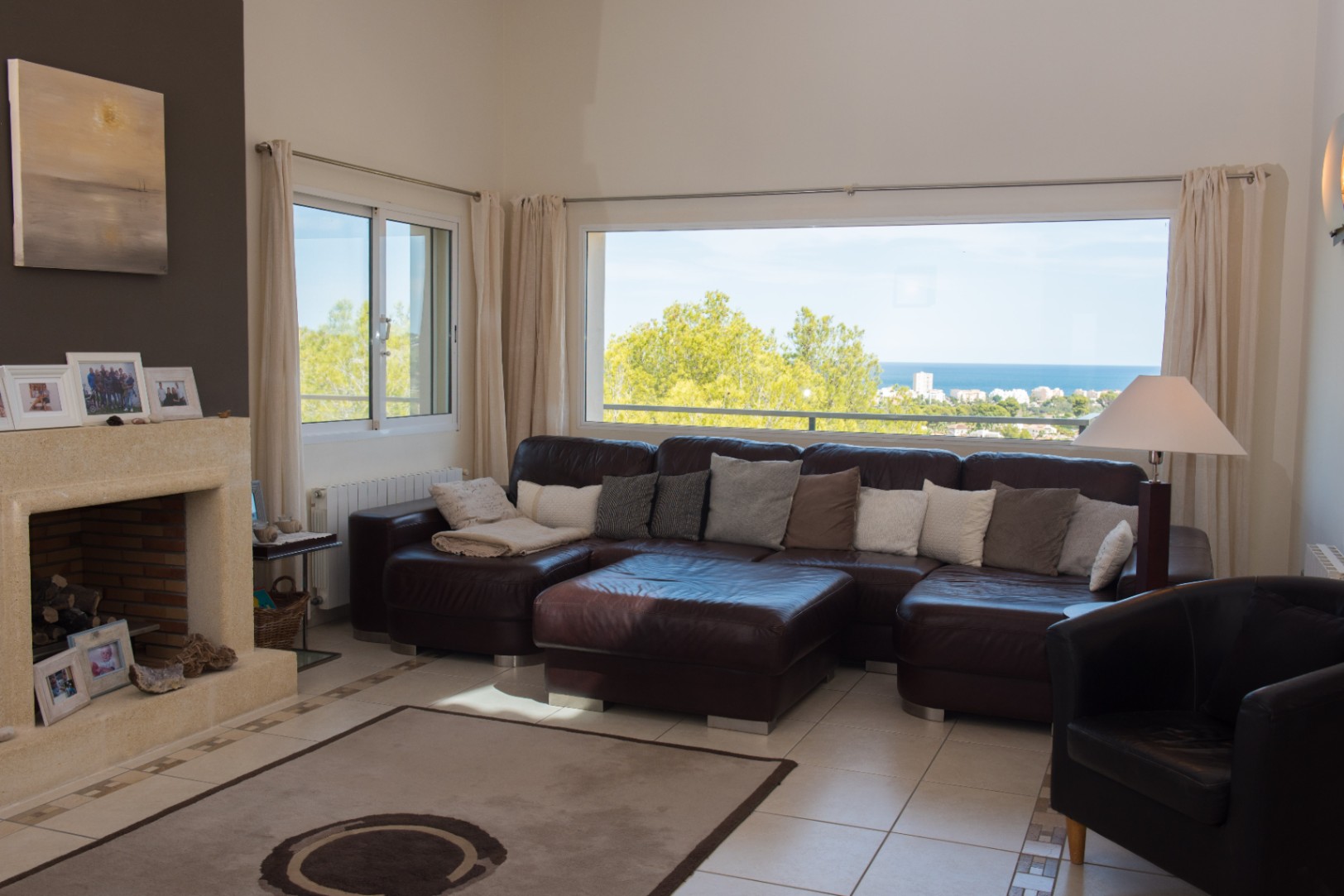 Amplio salón con sofá rinconero de cuero marrón, chimenea de piedra natural y un gran ventanal con vistas panorámicas al mar y la costa.