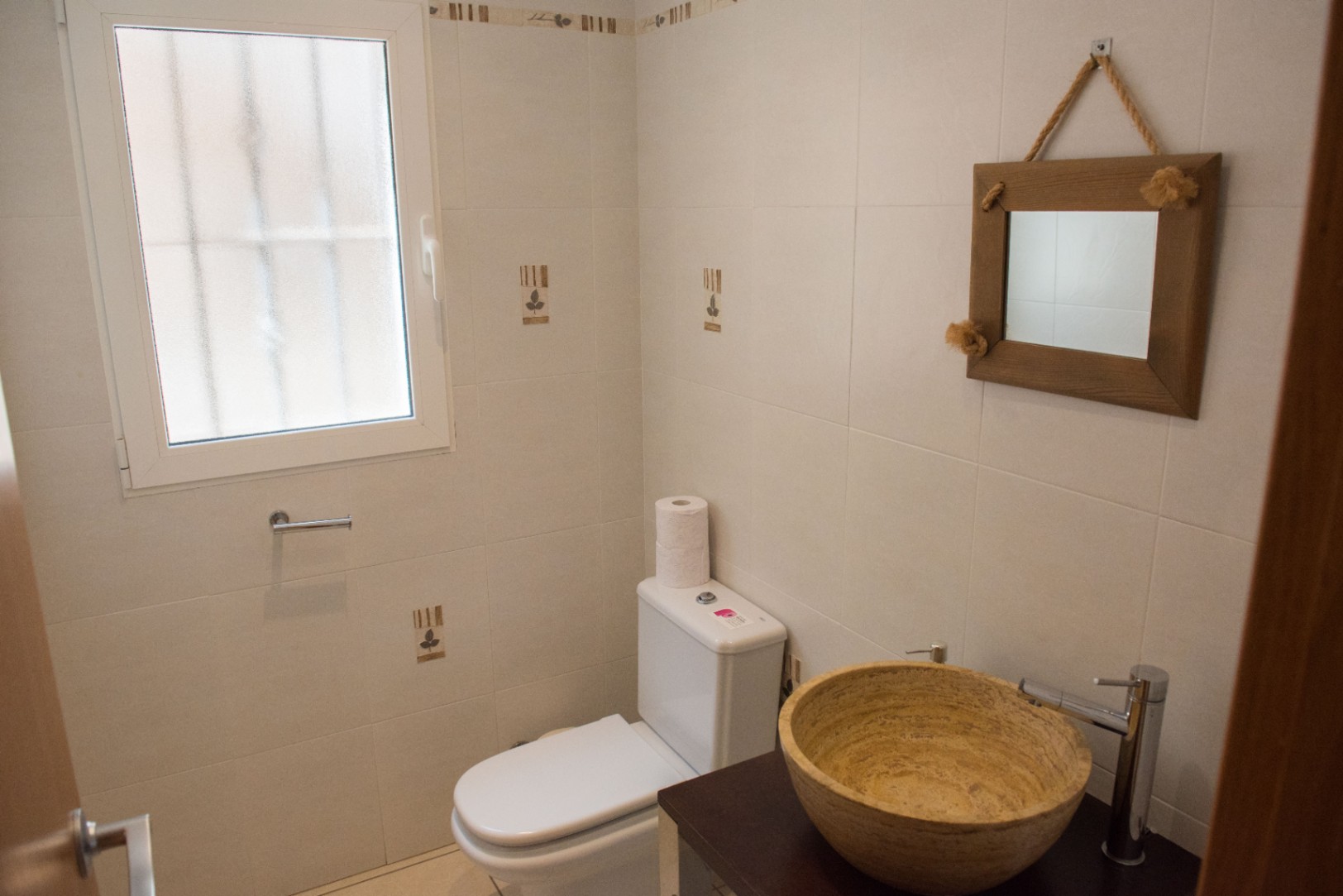 Aseo compacto con lavabo de piedra natural sobre encimera oscura, espejo con marco de madera y cuerda, y paredes alicatadas con detalles decorativos.