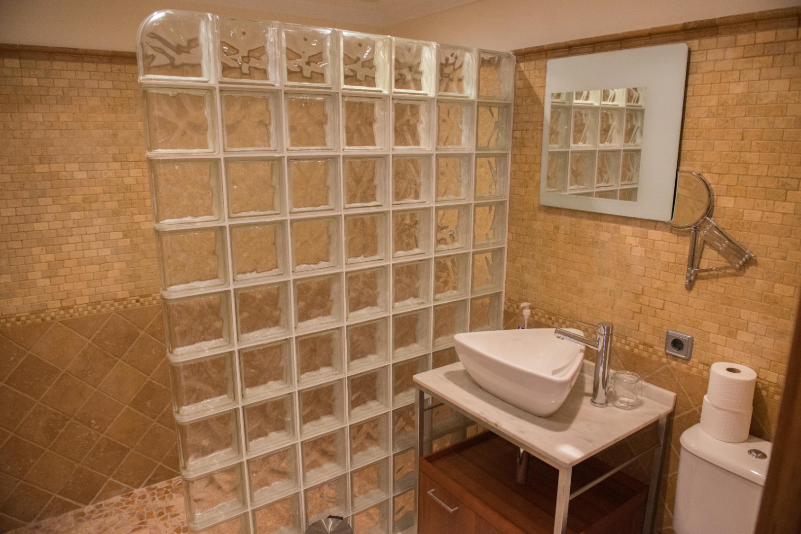 Baño con mampara de bloques de vidrio, paredes revestidas en mosaico beige y lavabo sobre encimera de mármol con mueble inferior de madera.