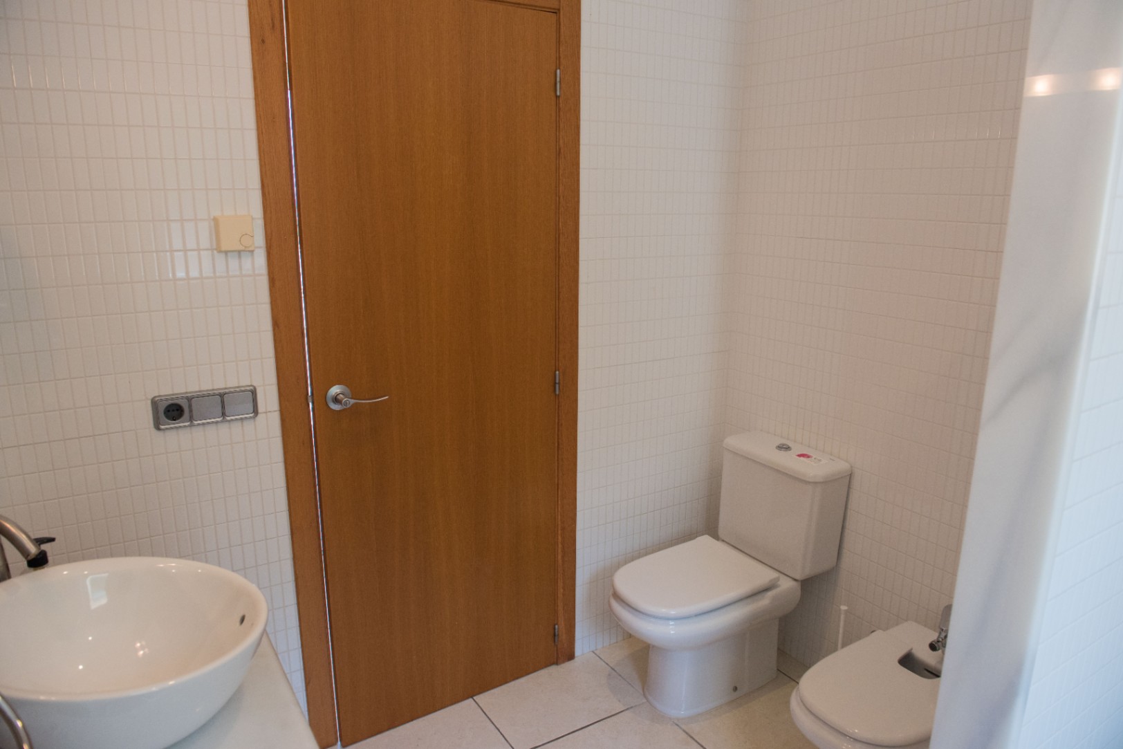 Baño con paredes de azulejos blancos, puerta de madera, inodoro de cerámica, bidé y un lavabo de sobremesa blanco con grifería cromada.
