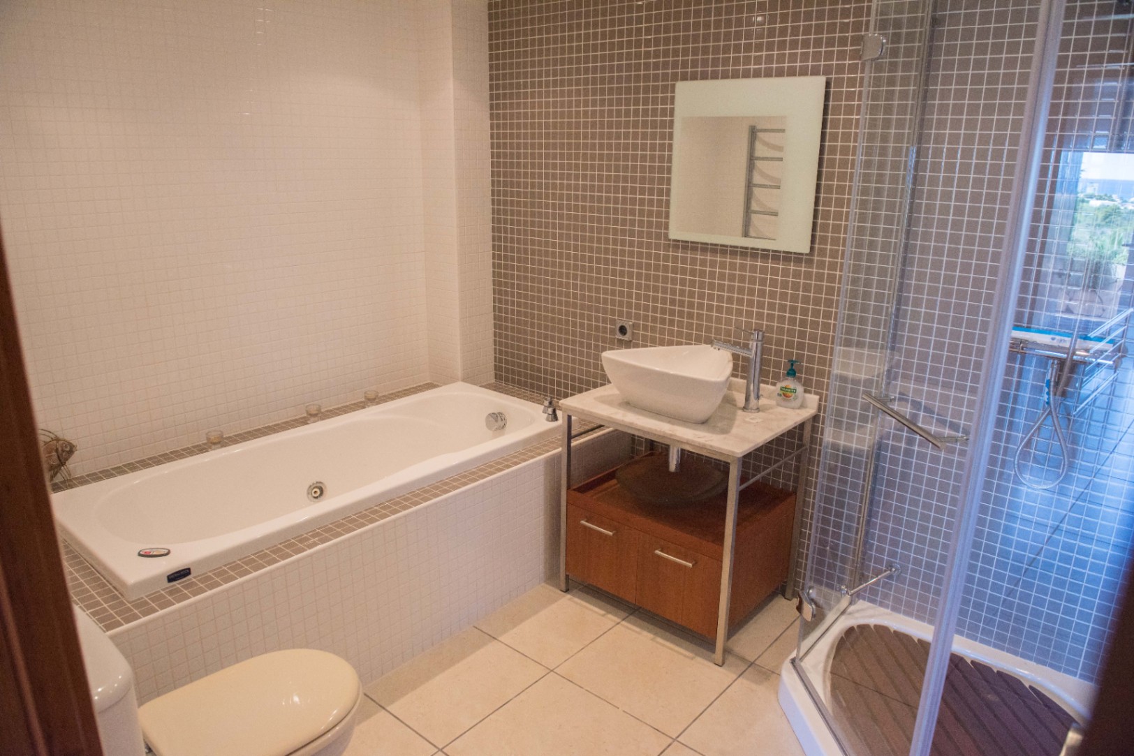 Baño moderno con bañera integrada, ducha de cristal con suelo de madera y lavabo sobre encimera de mármol, decorado con azulejos de mosaico en tonos neutros.