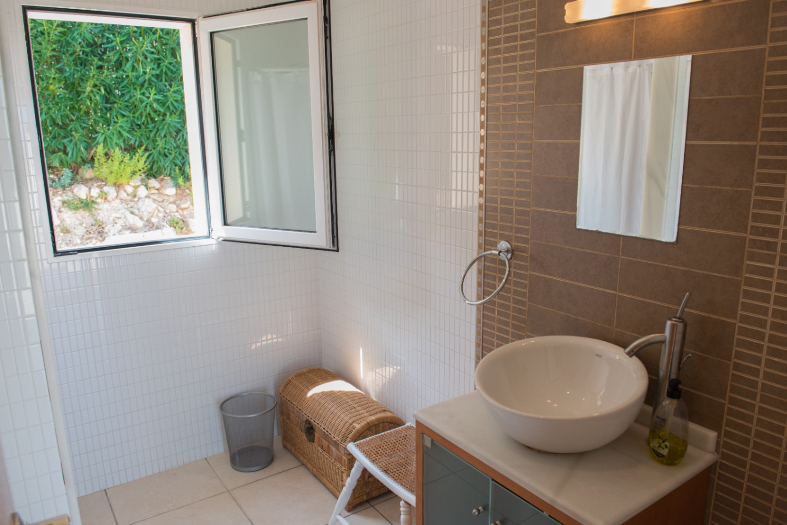 Baño moderno con lavabo de sobreponer, pared de acento en tonos marrones y ventana abierta con vistas al jardín exterior y luz natural.