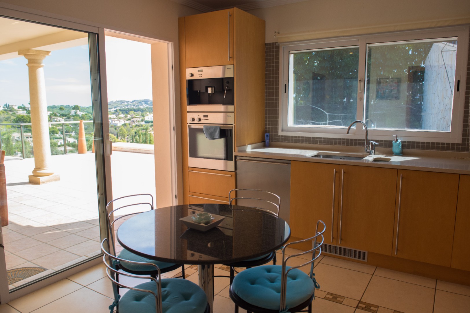 Cocina con armarios de madera clara, electrodomésticos Miele integrados y mesa de granito. El ventanal da acceso a una terraza con vistas panorámicas al valle.