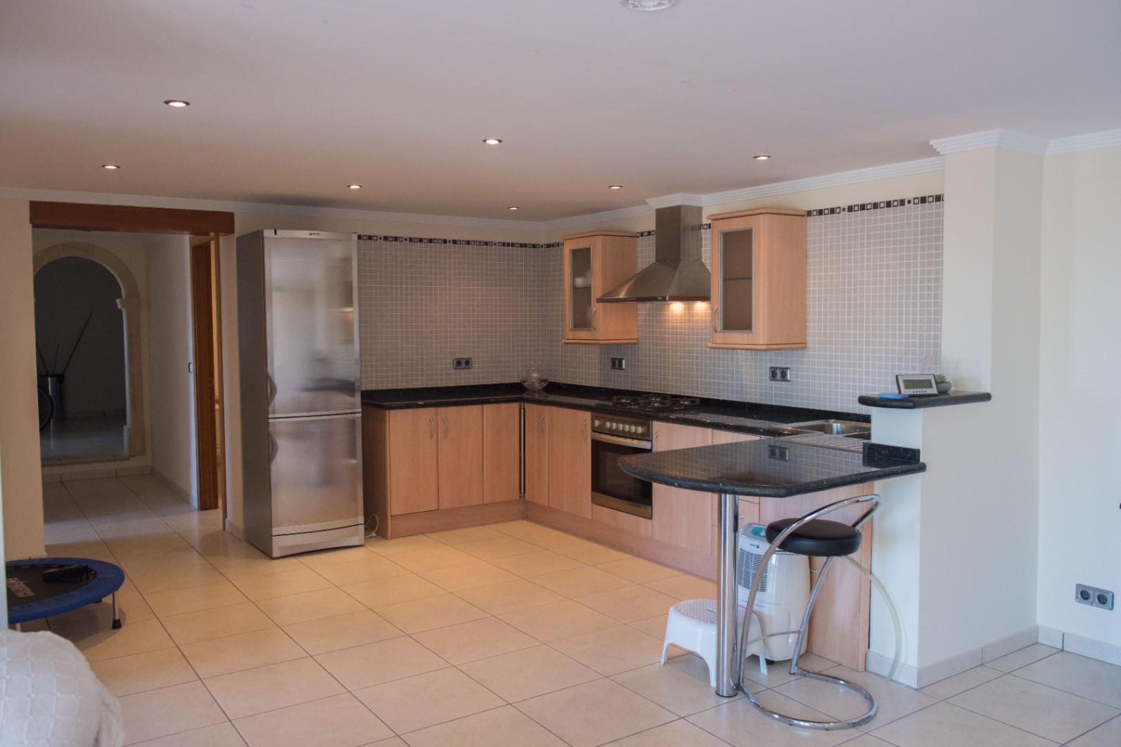 Cocina en L con armarios de madera clara, encimeras de granito oscuro y electrodomésticos de acero. Destaca el salpicadero de mosaico, la barra de desayuno y focos empotrados.