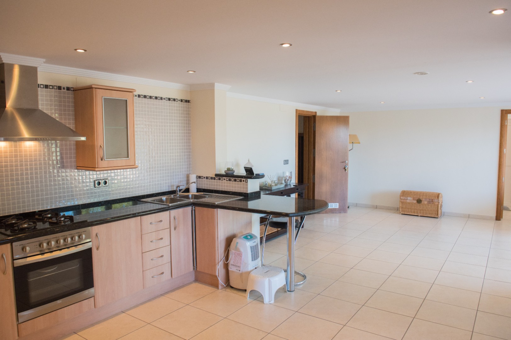 Cocina funcional en forma de L con armarios de madera clara, encimeras de granito oscuro y barra americana. Incluye electrodomésticos de acero inoxidable y focos empotrados.