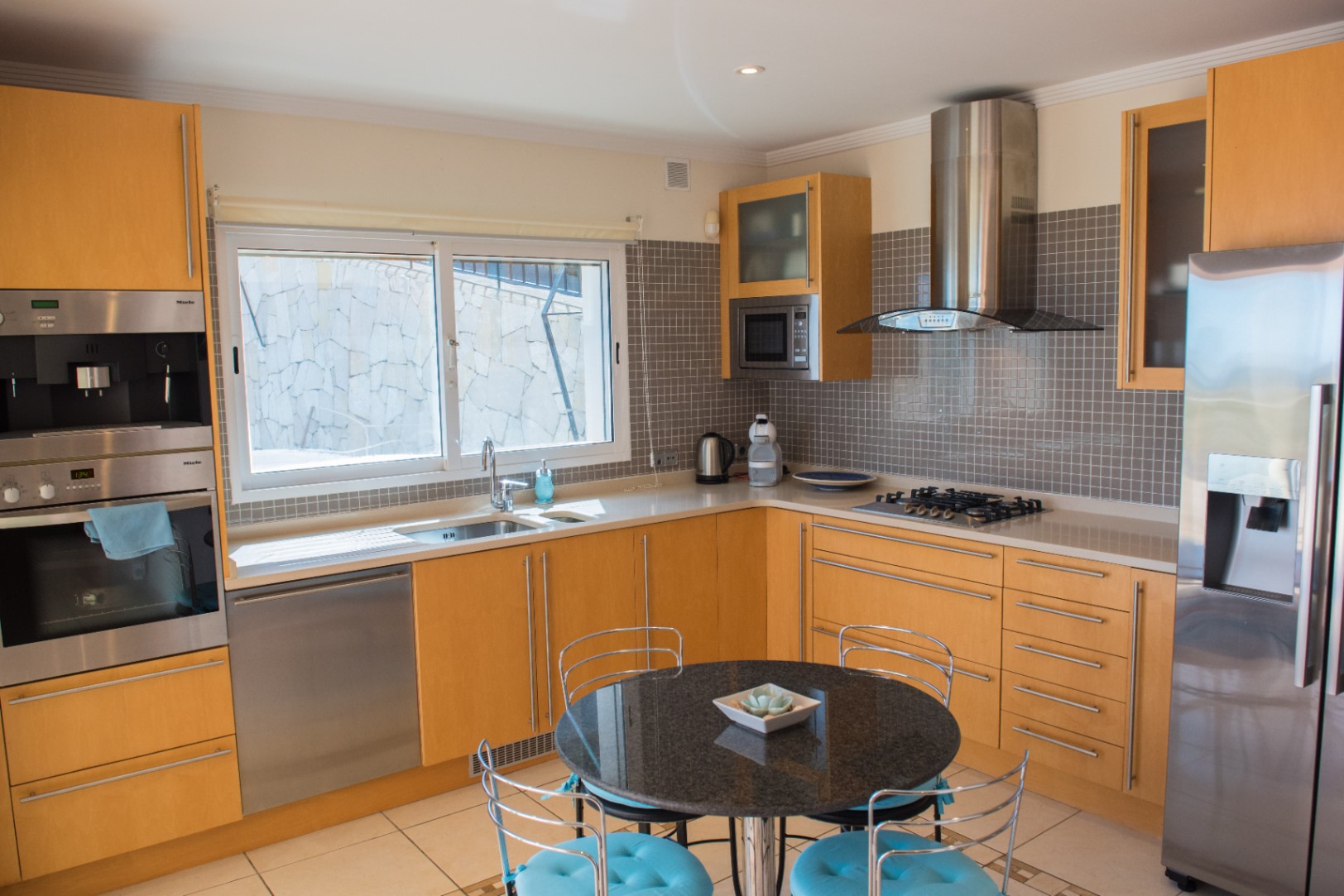 Cocina moderna con armarios de madera clara, electrodomésticos Miele de acero inoxidable y salpicadero de mosaico gris. Incluye una mesa de granito negro y encimeras blancas.