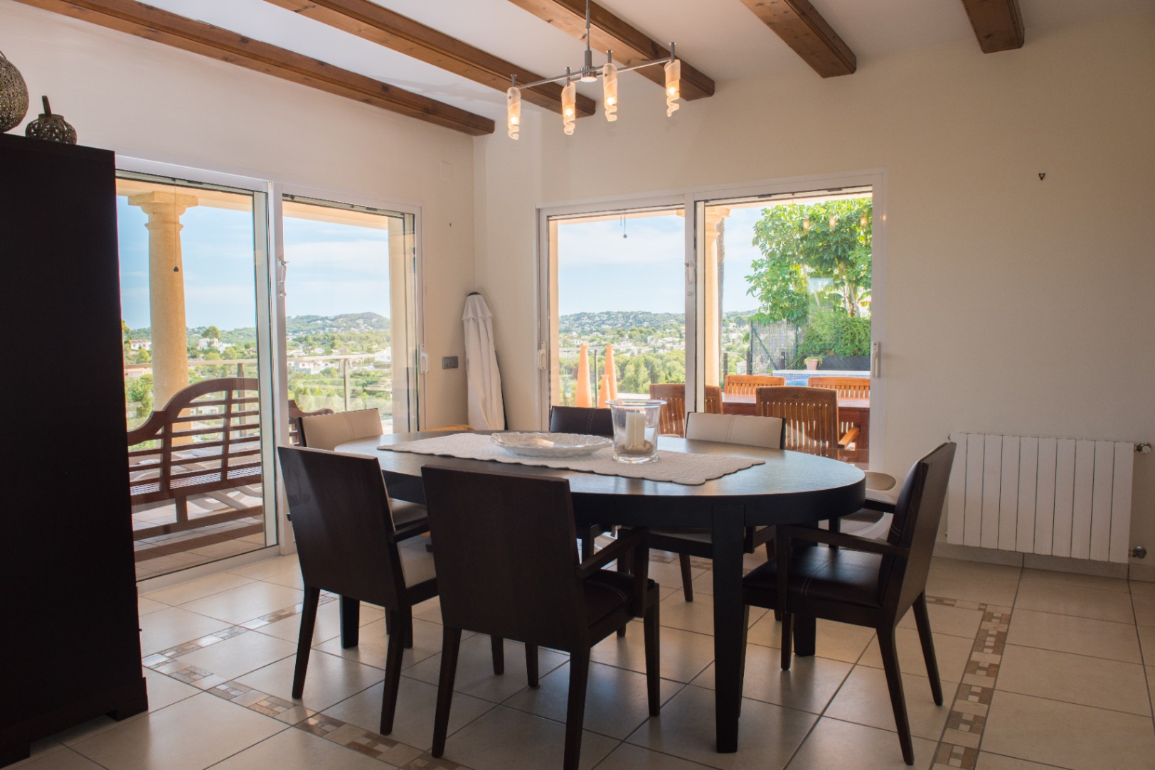 Comedor con mesa ovalada de madera oscura, vigas de madera vistas y amplios ventanales correderos que ofrecen vistas panorámicas a las colinas y acceso a la terraza.
