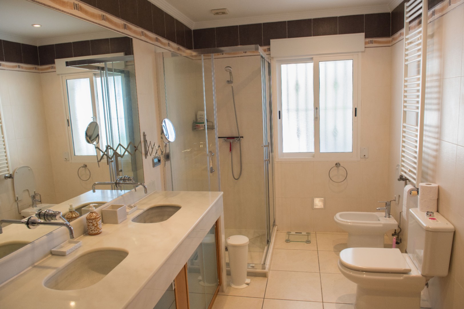 Cuarto de baño con lavabo doble de mármol, cabina de ducha acristalada y sanitarios blancos, complementado por un gran espejo y azulejos en tonos neutros.