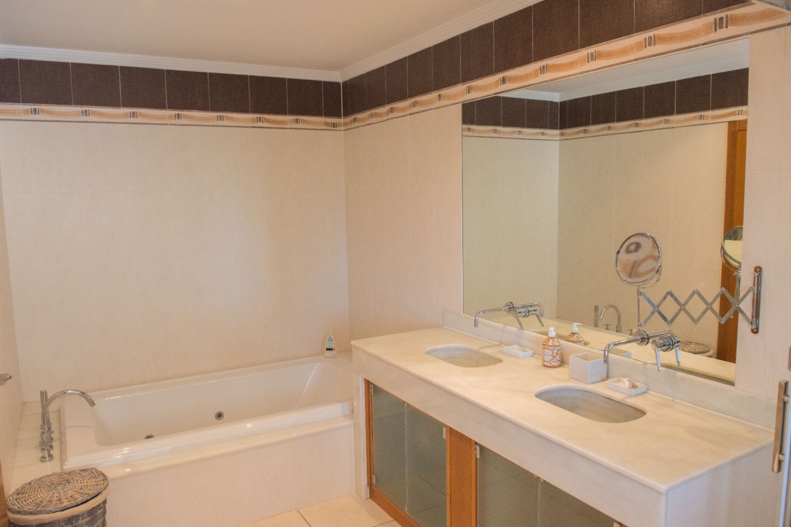 Cuarto de baño con lavabo doble de mármol, grifería de pared y bañera de hidromasaje integrada, decorado con azulejos en tonos crema y cenefa oscura.