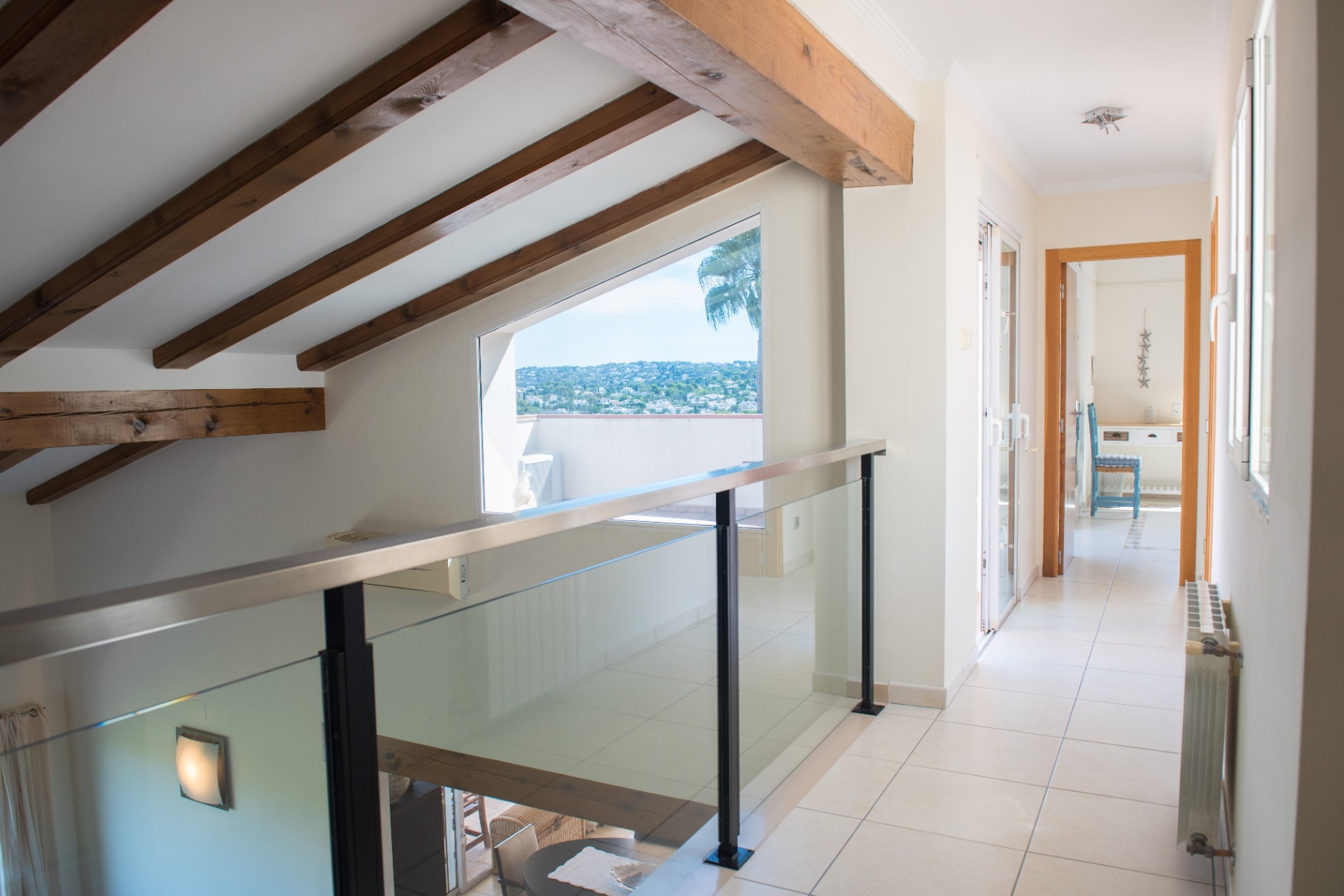 Distribuidor en planta superior con vigas de madera a la vista, barandilla de cristal y un gran ventanal trapezoidal con vistas panorámicas al entorno.
