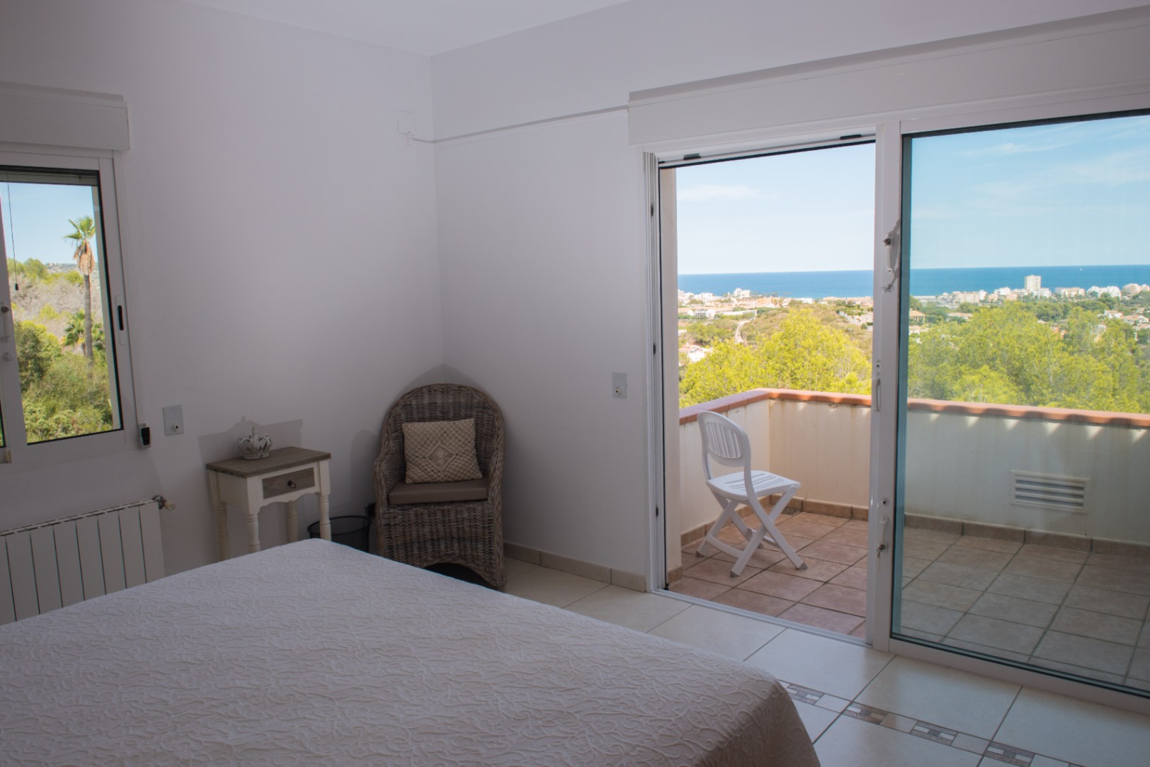 Dormitorio con acceso a balcón privado y vistas panorámicas al mar. Cuenta con suelos cerámicos claros, un sillón de mimbre y carpintería exterior de aluminio blanco.