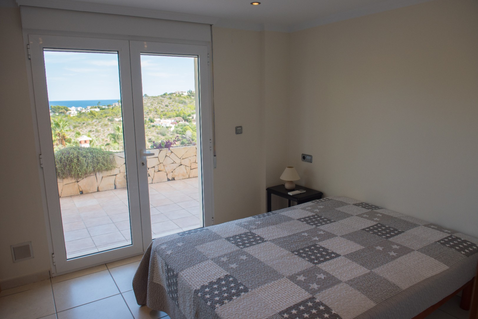 Dormitorio con acceso directo a una terraza privada mediante puertas acristaladas, con vistas al mar y al entorno natural. Incluye suelos cerámicos y abundante luz natural.