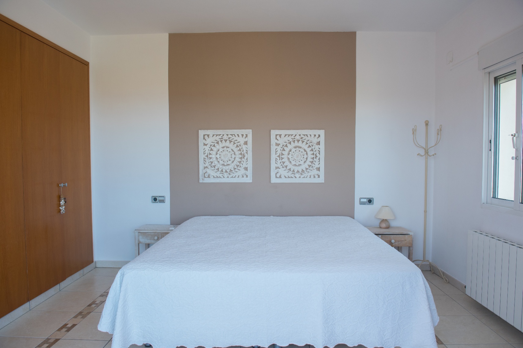 Dormitorio con pared de acento en tono topo, paneles decorativos tallados, armario empotrado de madera y suelo cerámico con cenefa decorativa.