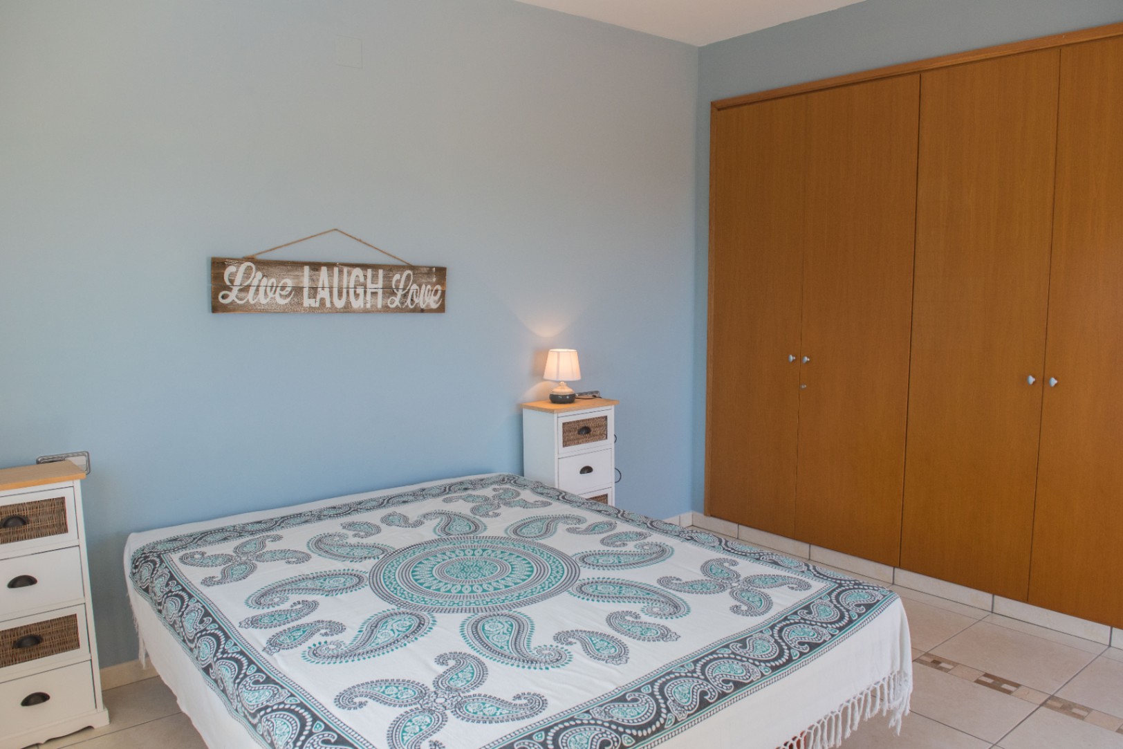Dormitorio con paredes azul claro, un amplio armario empotrado de madera y muebles blancos con detalles de mimbre. Presenta suelos cerámicos y una colcha estampada.