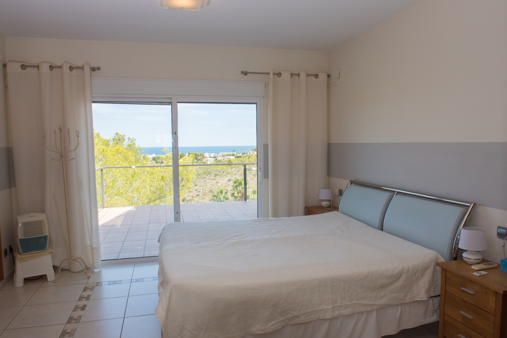 Dormitorio con suelos cerámicos y puertas correderas hacia una terraza con vistas al mar. Destaca su cabecero metálico moderno y paredes en tonos neutros.