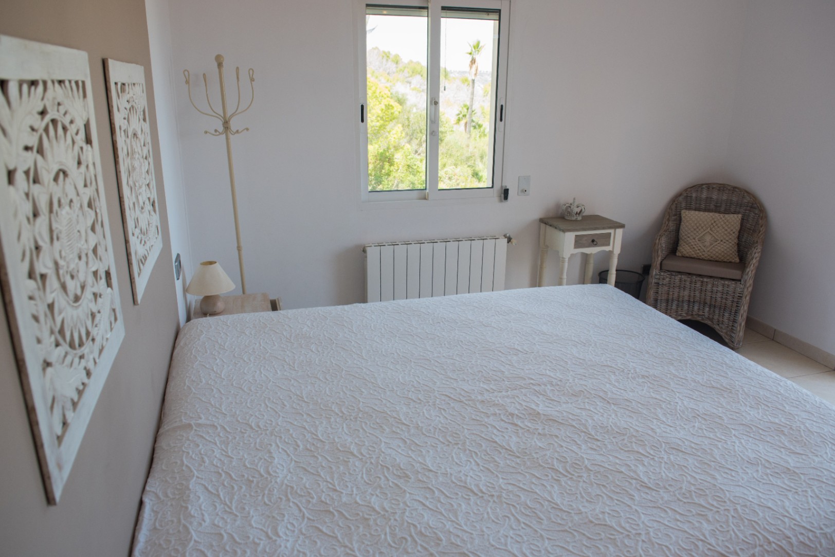 Dormitorio luminoso con colcha blanca texturizada, paneles de madera tallada y un sillón de mimbre. La ventana ofrece vistas a la vegetación exterior y palmeras.