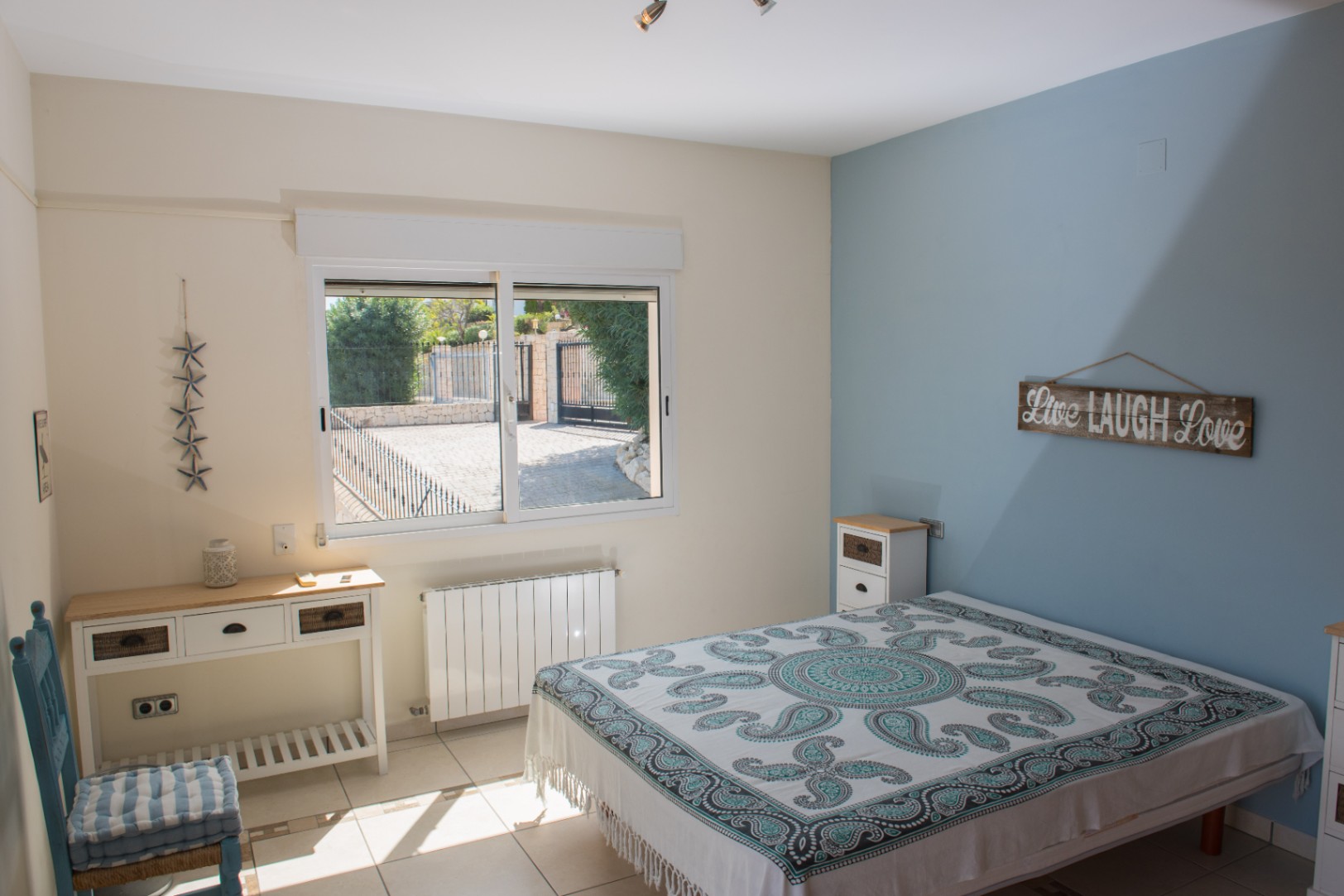 Dormitorio luminoso con pared de acento azul, suelos cerámicos y ventana con vistas al patio exterior vallado. Incluye escritorio blanco y radiador de pared.