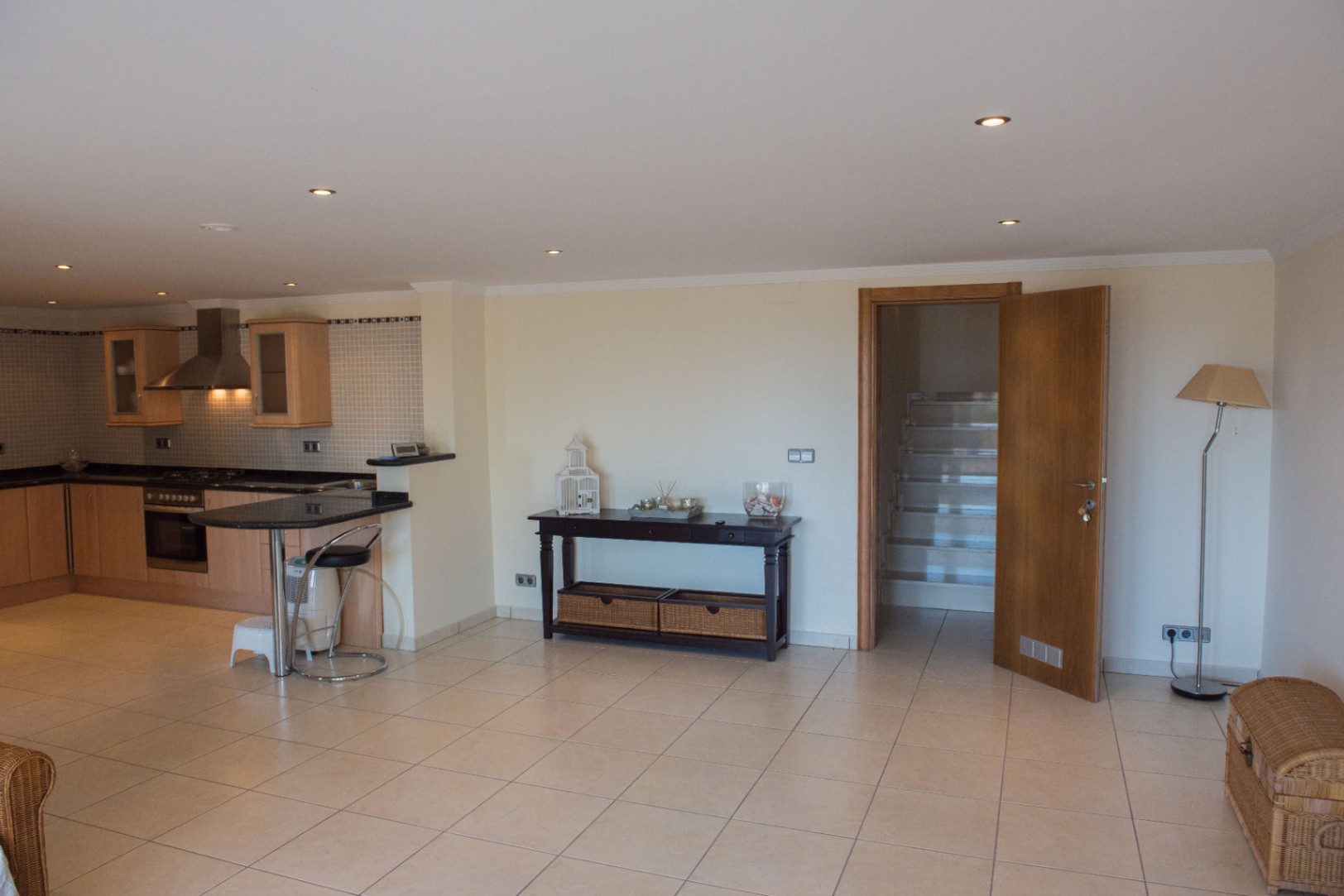 Espacio diáfano con cocina de armarios de madera clara, encimeras de granito negro y barra americana, junto a un recibidor con suelos cerámicos y acceso a la escalera.