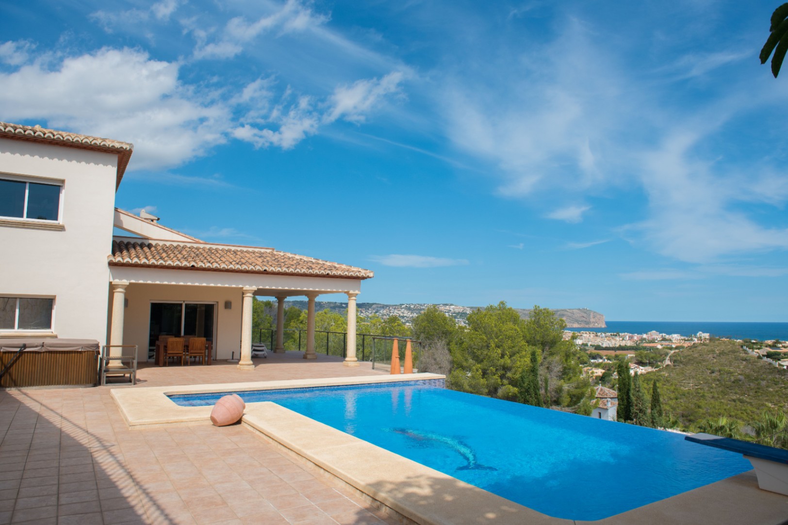 Piscina infinita con mosaico de delfín y vistas panorámicas al mar. La villa dispone de un porche cubierto con columnas de piedra y tejado de teja cerámica.