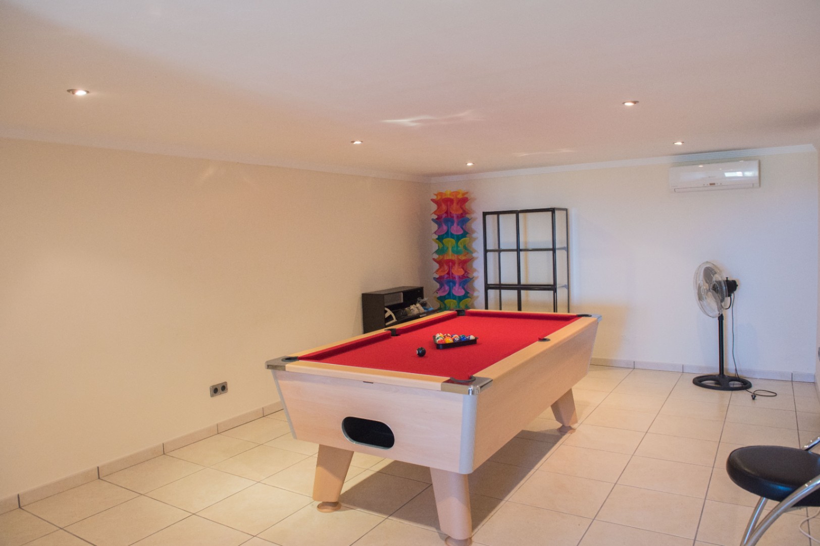 Sala de juegos con mesa de billar de tapete rojo y estructura de madera clara, suelo cerámico, iluminación empotrada y aire acondicionado.