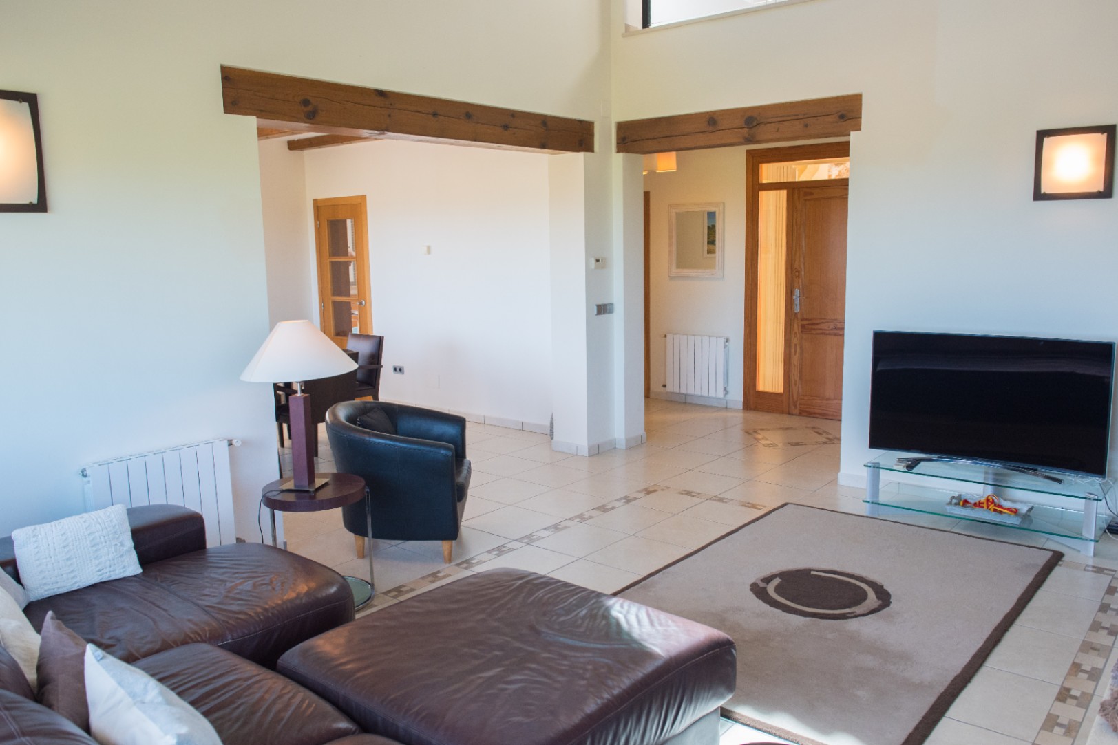 Salón con sofás de cuero oscuro, suelos cerámicos con cenefas y vigas de madera a la vista. El diseño abierto conecta con el comedor y puertas de madera natural.