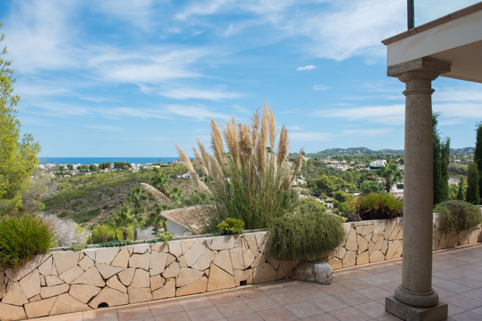 Terraza con columnas de piedra y muro perimetral de mampostería, decorada con plumeros y vistas panorámicas a la costa mediterránea y colinas circundantes.