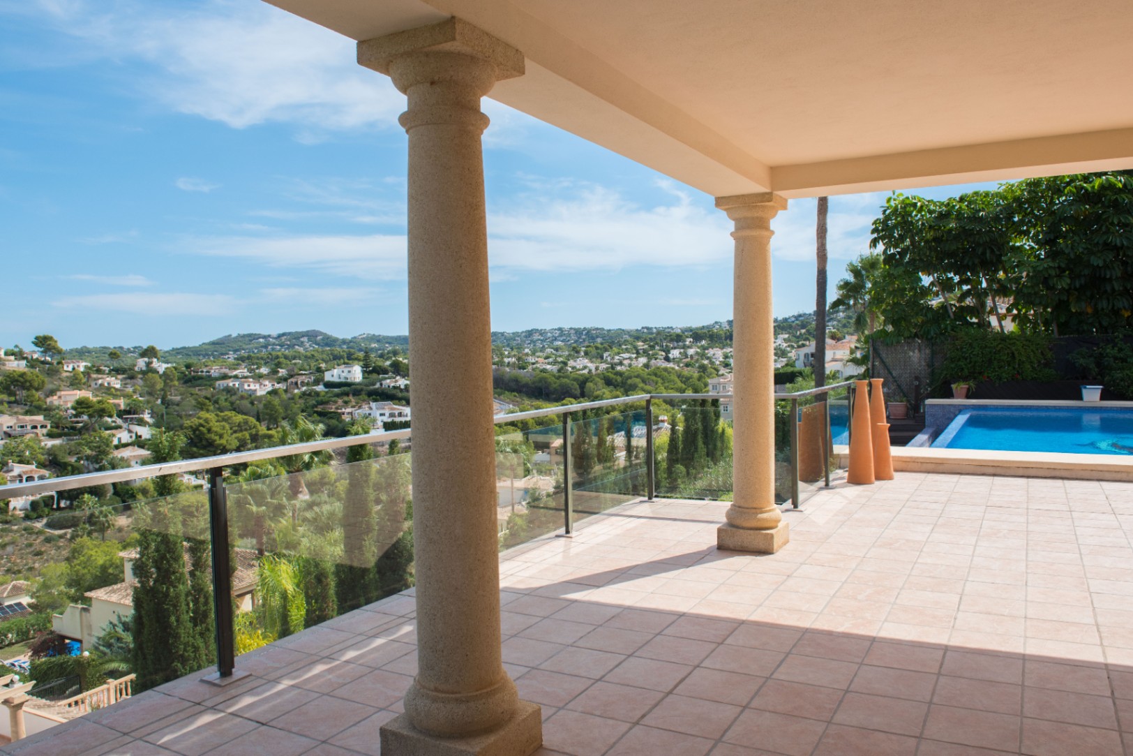 Terraza cubierta con columnas de piedra y barandilla de cristal, con vistas panorámicas al valle y acceso a la piscina.