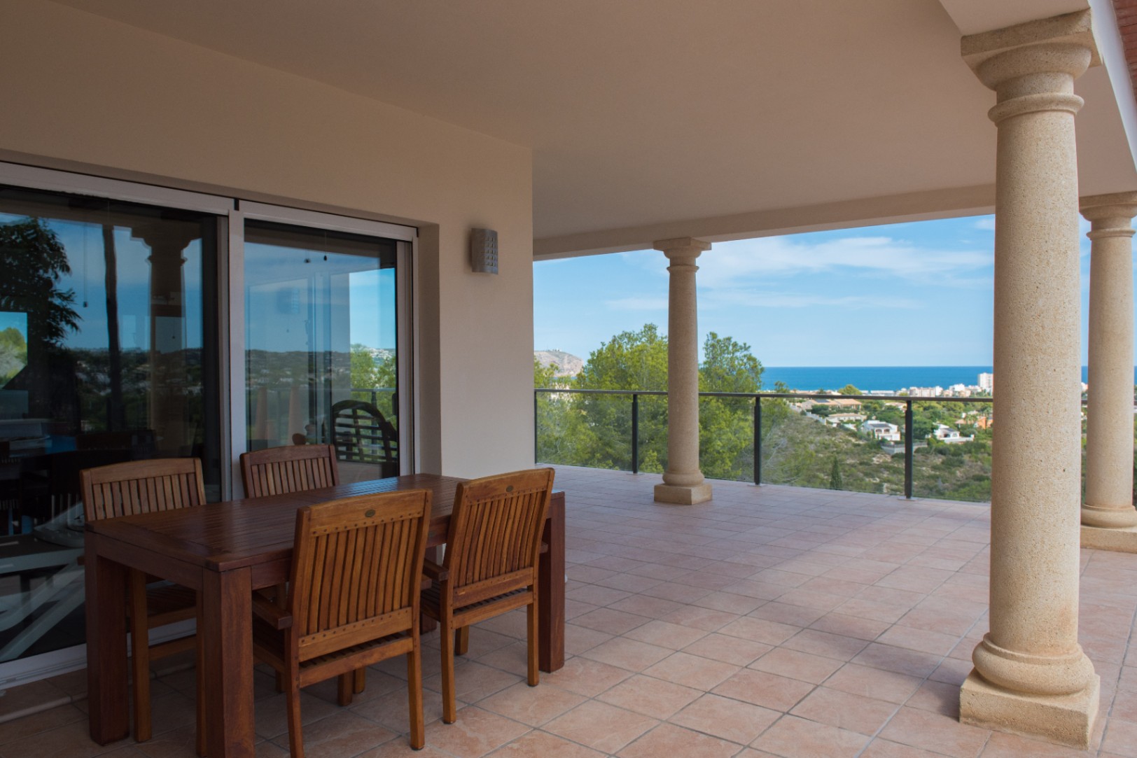 Terraza cubierta con mesa de comedor de madera, columnas de piedra de estilo clásico y vistas panorámicas al mar tras una barandilla de cristal.