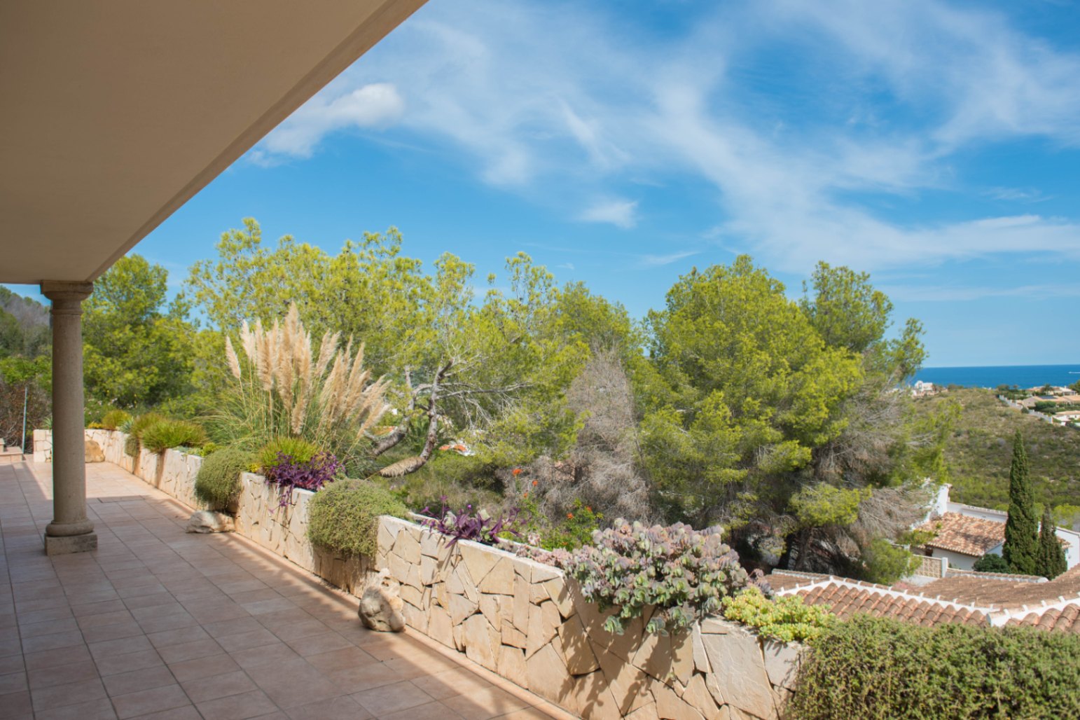 Terraza cubierta con suelos cerámicos y jardinera de piedra natural con vegetación mediterránea, vistas despejadas a los pinos y al mar.