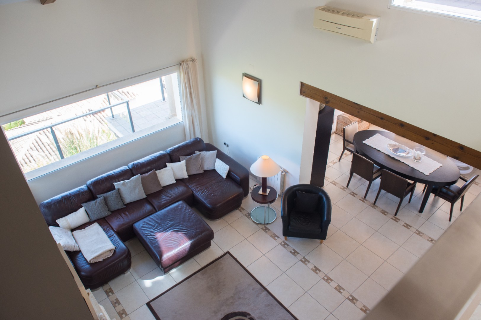 Vista elevada de un salón con sofá rinconero de cuero marrón, suelos cerámicos con cenefas y un comedor contiguo con mesa ovalada bajo una viga de madera vista.