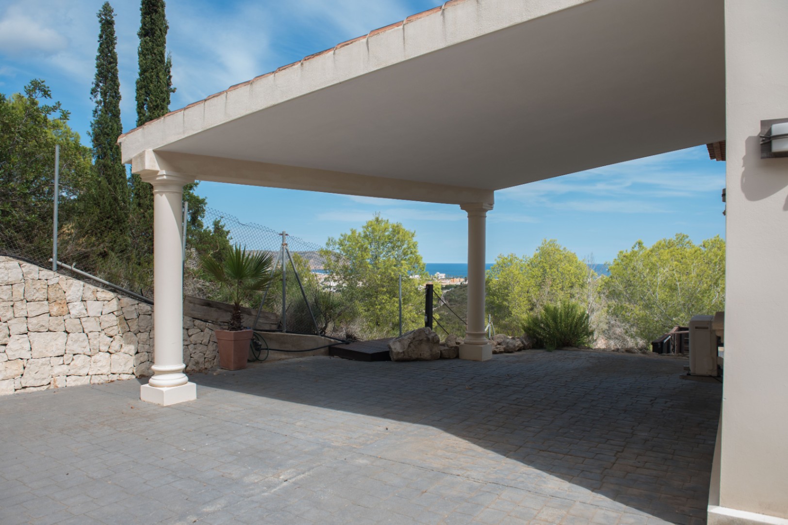 Zona de aparcamiento cubierta con columnas clásicas y suelo de adoquines, con vistas despejadas al mar Mediterráneo y vegetación autóctona.